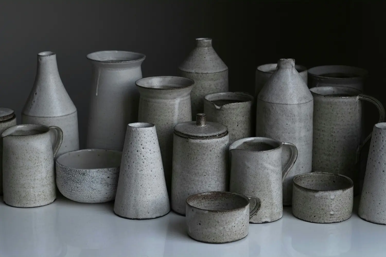 Mesa del taller con jarras, botellas y cuencos de gres blanco moteado sobre fondo gris oscuro